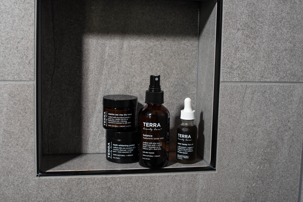 Terra Beauty Bars products in bathroom shelf to include matcha sea clay dry mask in glass jar 2oz, teeth whitening polish in black plastic  jar 2oz, Balance hyaluronic toner mist in amber 4 oz glass bottle spray and 1 oz hemp oil in frosted glass package